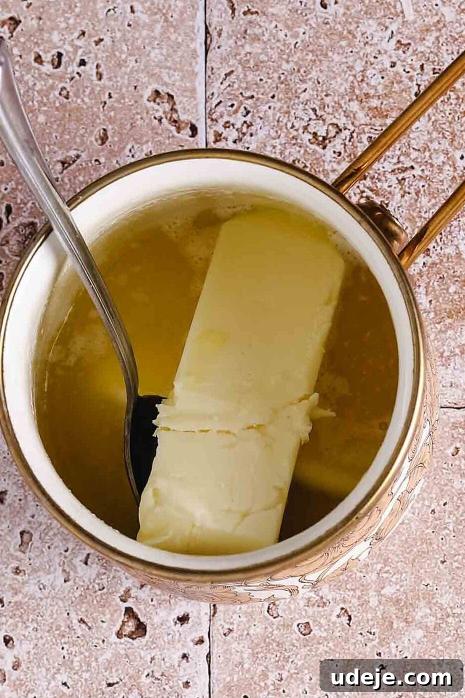 A small gold saucepan with a stick of butter and melted butter, with a spoon sticking out, on a light stone tiled background.