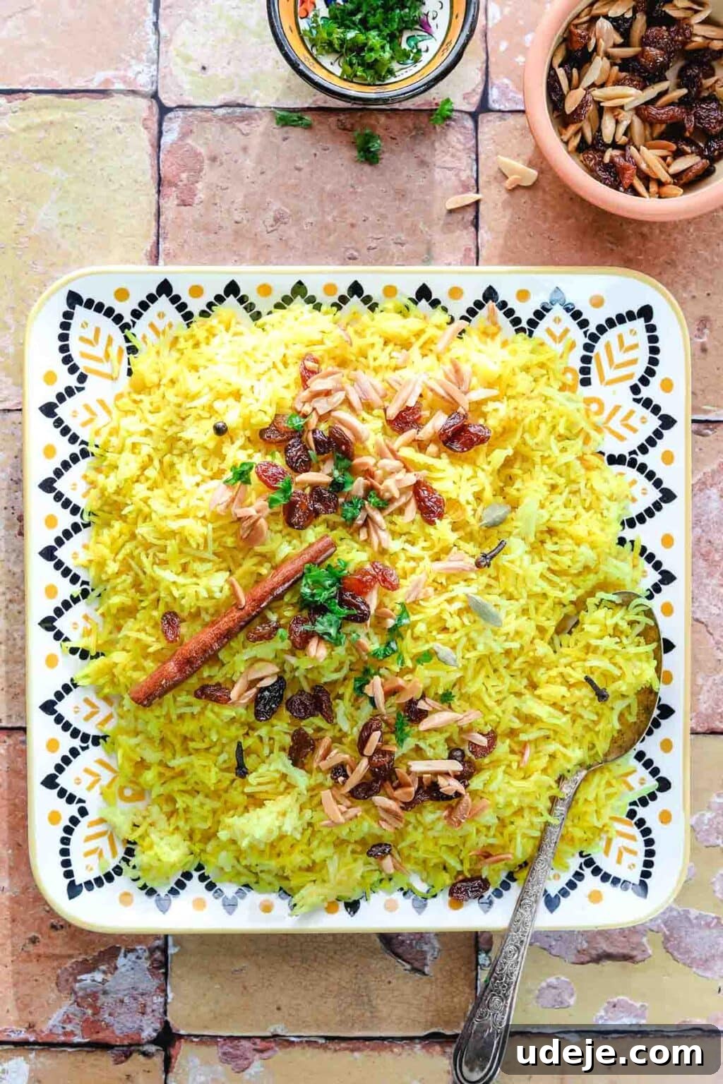 30-Minute Golden Mediterranean Basmati 2 A plate of vibrant Mediterranean Yellow Rice garnished with golden raisins, delicate slivered almonds, fresh herbs, and a decorative cinnamon stick. A patterned spoon rests gracefully on the plate, with small accompanying bowls of extra toppings arranged on a beautifully patterned tile surface, inviting a taste of this aromatic dish.