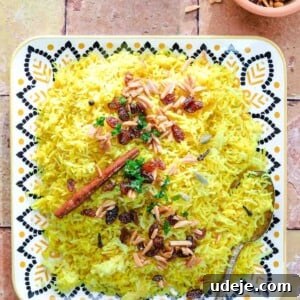 A decorative plate of Mediterranean Yellow Rice, filled with saffron rice, raisins, slivered almonds, chopped herbs, and a cinnamon stick. A spoon rests in the rice, with small side bowls of toppings nearby on a tiled surface.