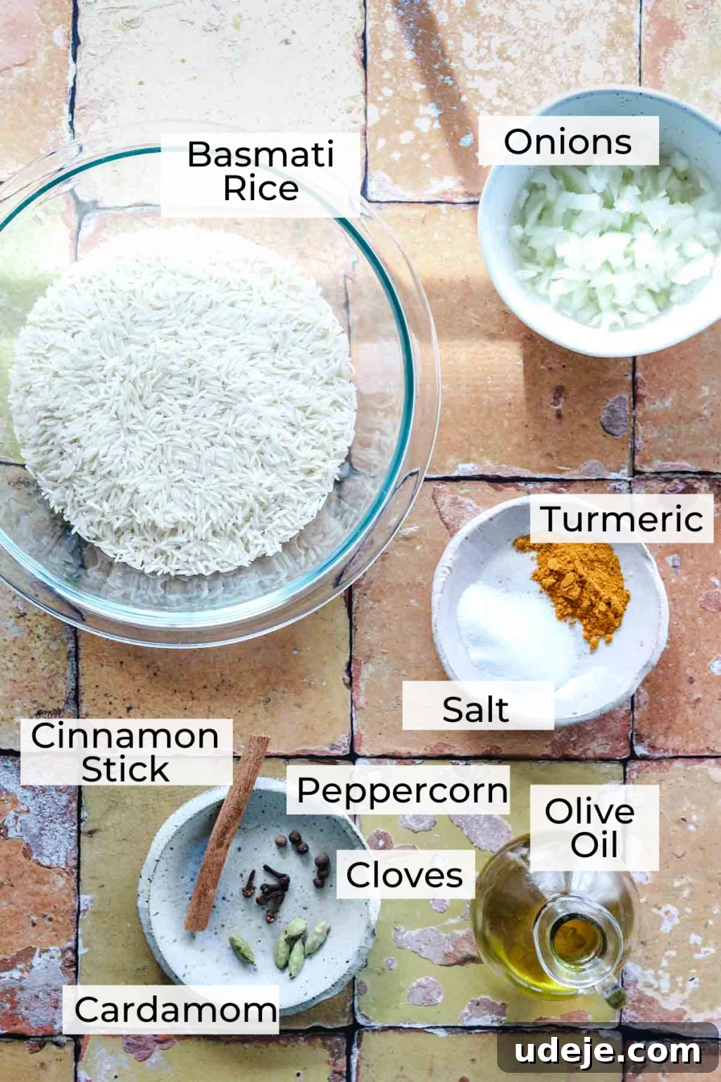 30-Minute Golden Mediterranean Basmati 3 A visually appealing flat lay of Mediterranean Yellow Rice ingredients spread across a tiled surface. Key components include mounds of uncooked basmati rice, finely chopped onions, vibrant turmeric powder, coarse salt, and a bottle of golden olive oil. Small individual bowls display whole spices such as a fragrant cinnamon stick, glossy black peppercorns, aromatic cloves, and delicate green cardamom pods. Each ingredient is thoughtfully arranged and labeled, showcasing the fresh and whole nature of the recipe's components.