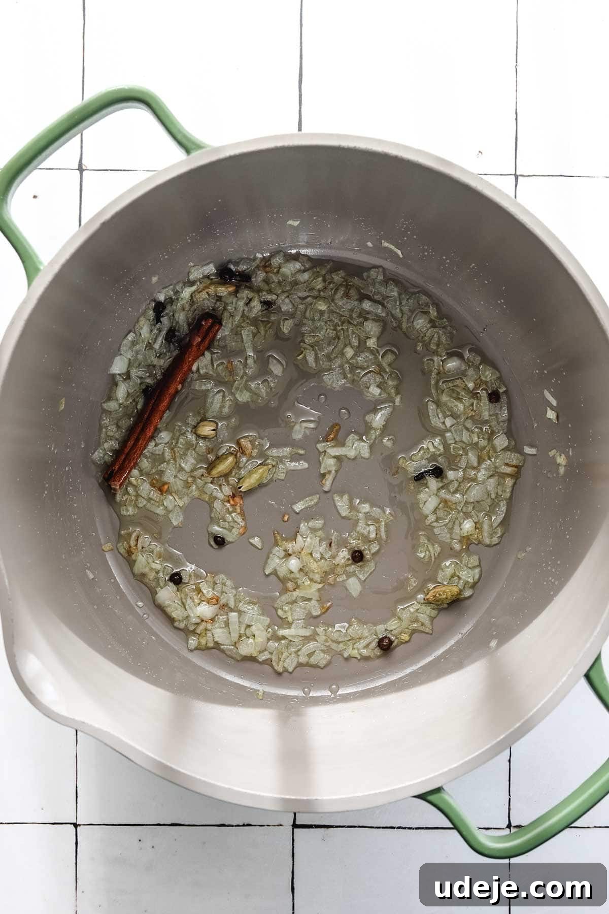 30-Minute Golden Mediterranean Basmati 4 A large cooking pot sits on a white tiled surface, filled with a fragrant mixture of golden sautéed chopped onions, glistening in oil. Amidst the onions, whole spices are visible, including a prominent cinnamon stick, small dark cloves, and scattered peppercorns, all gently cooking to form the aromatic base for Mediterranean Yellow Rice. The subtle steam rising indicates the beginning of a flavorful culinary process.