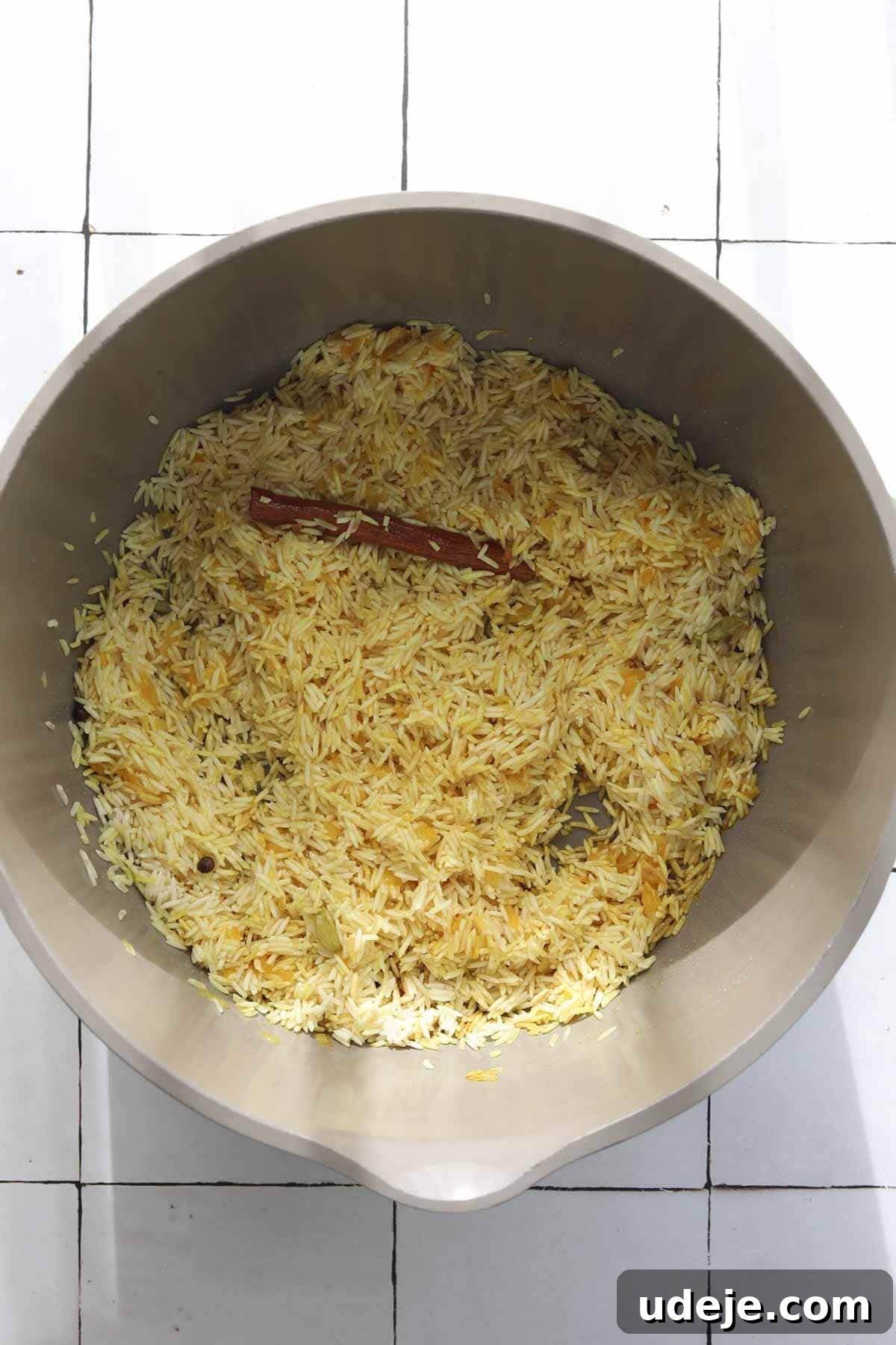 30-Minute Golden Mediterranean Basmati 6 A metal mixing bowl holds dry, uncooked basmati rice, now infused with the golden hue of turmeric and mixed with whole spices, including a prominent cinnamon stick. The grains appear distinct and ready for the next stage of cooking, resting on a clean white tiled surface. This close-up highlights the prepared rice before liquid is added, showcasing its vibrant color and aromatic potential.