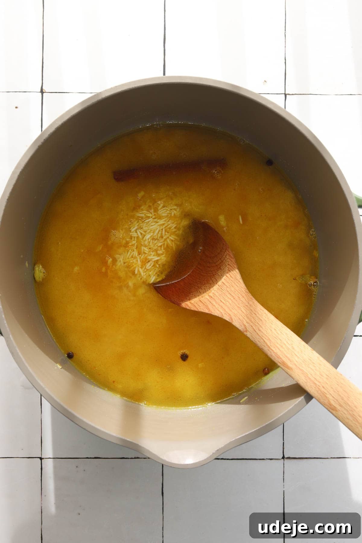 30-Minute Golden Mediterranean Basmati 7 A pot of Mediterranean Yellow Rice ingredients, including golden liquid, basmati rice, and whole spices such as a visible cinnamon stick, is being stirred with a wooden spoon on a white tiled surface. The liquid is just beginning to simmer, indicating the rice is absorbing the rich flavors. The image captures the crucial moment before the pot is covered for cooking, showcasing the blend of ingredients and the start of the cooking process.
