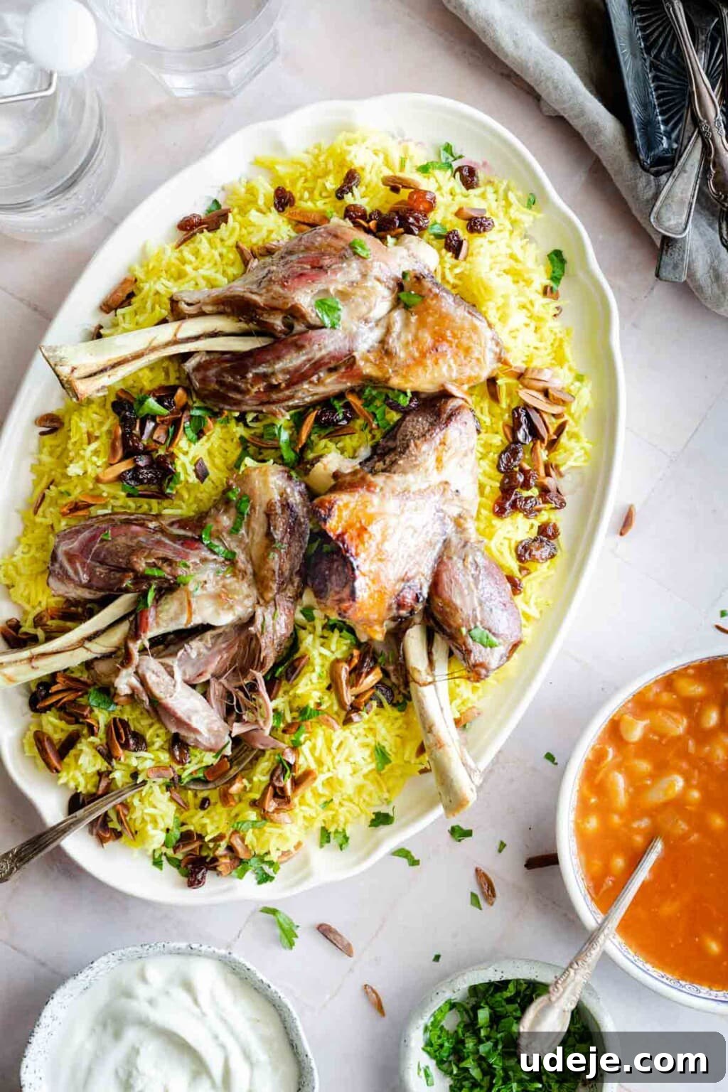 30-Minute Golden Mediterranean Basmati 9 A grand platter showcasing Quzi-style cooked lamb shanks, tender and roasted to perfection, elegantly served over a generous bed of vibrant yellow rice. The rice is richly garnished with sweet raisins, fresh green herbs, and toasted nuts, adding layers of flavor and texture. Accompanying the main dish are several small, inviting bowls containing creamy yogurt, a rich tomato stew, extra chopped herbs, and essential cutlery and salt shakers, all contributing to a complete and delightful Middle Eastern feast.
