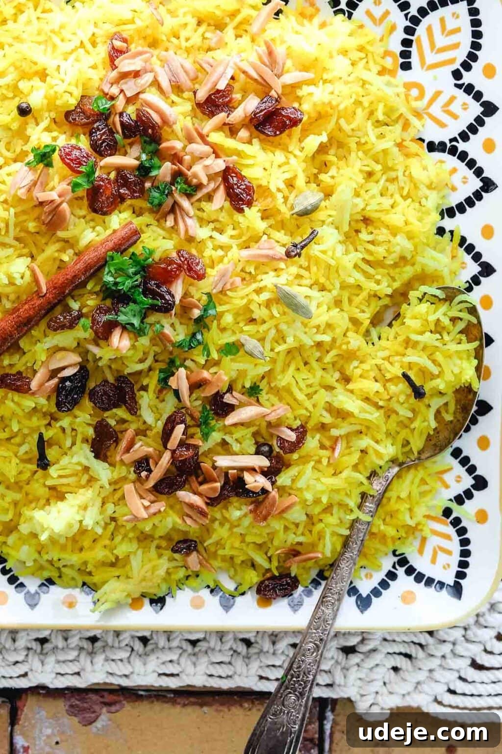 30-Minute Golden Mediterranean Basmati 10 A visually stunning plate of Mediterranean Yellow Rice, perfectly cooked and glistening. The rice is artfully garnished with crunchy slivered almonds, sweet golden raisins, and vibrant fresh parsley. Whole spices, including a cinnamon stick, are subtly visible, hinting at the aromatic depth of the dish. A decorative patterned spoon rests invitingly on the side of the plate, all set on a light-colored surface. This presentation emphasizes the versatility and rich flavor profile of this savory yellow rice.