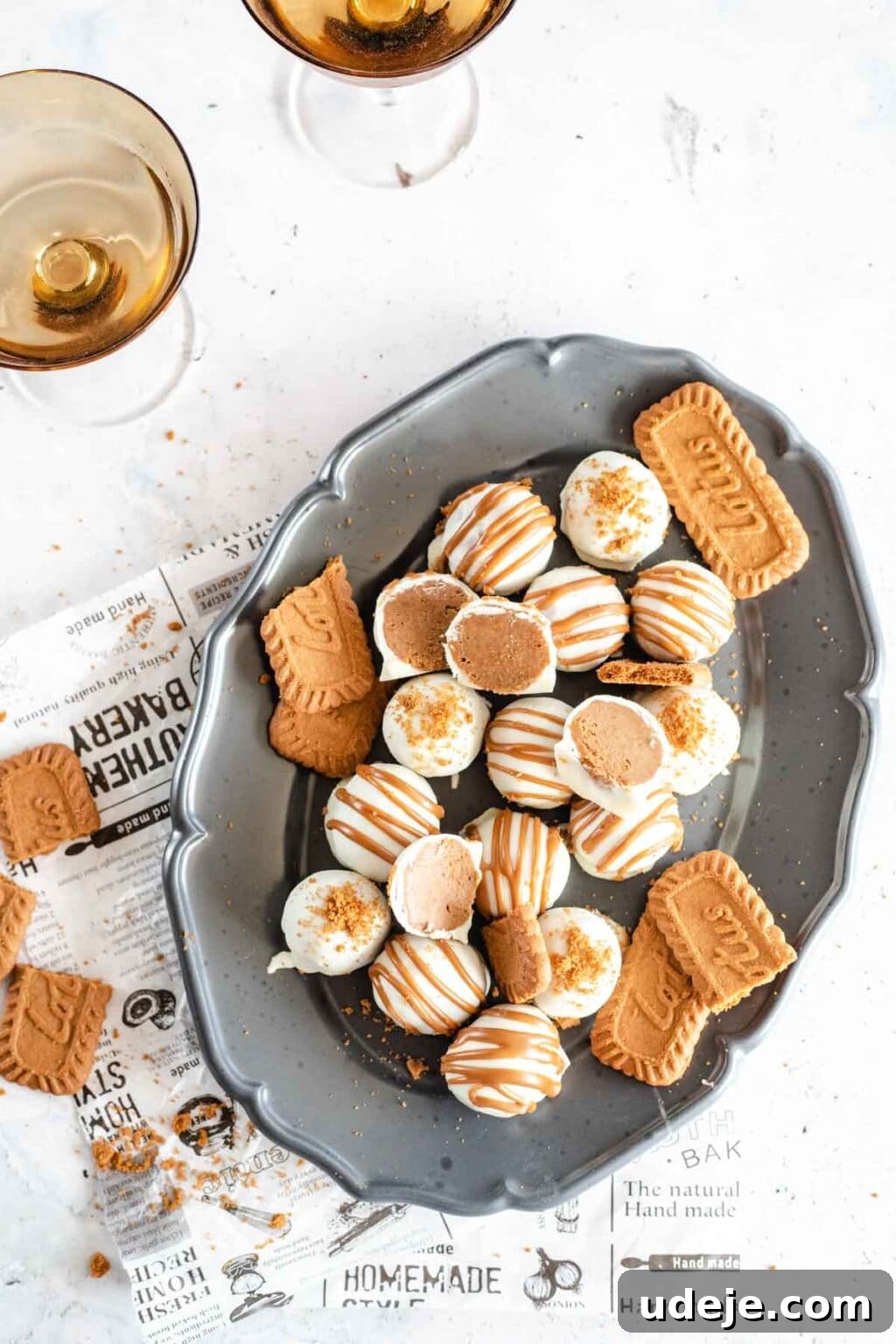4-Ingredient No-Bake Biscoff Truffles 2 Decadent Biscoff truffles elegantly arranged on a gray oval plate, resting on a newspaper sheet, with amber-colored glasses in the background against a pristine white marble surface.