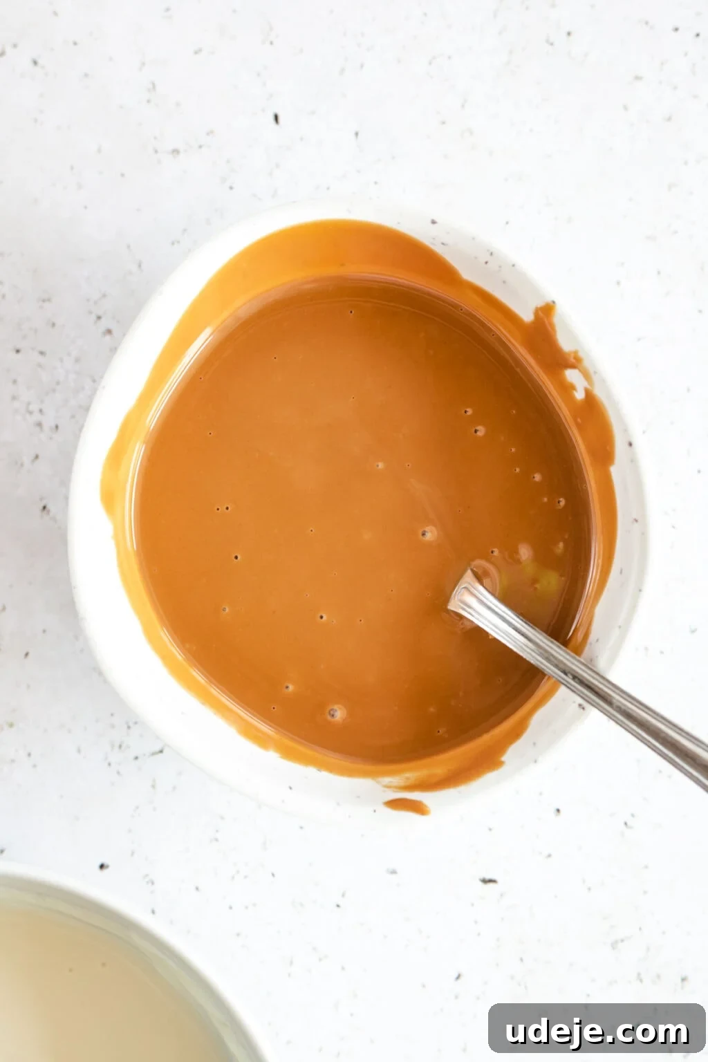 4-Ingredient No-Bake Biscoff Truffles 12 Melted Biscoff spread in a white bowl with a spoon, positioned alongside melted white chocolate in another white bowl, on a light marbled background.