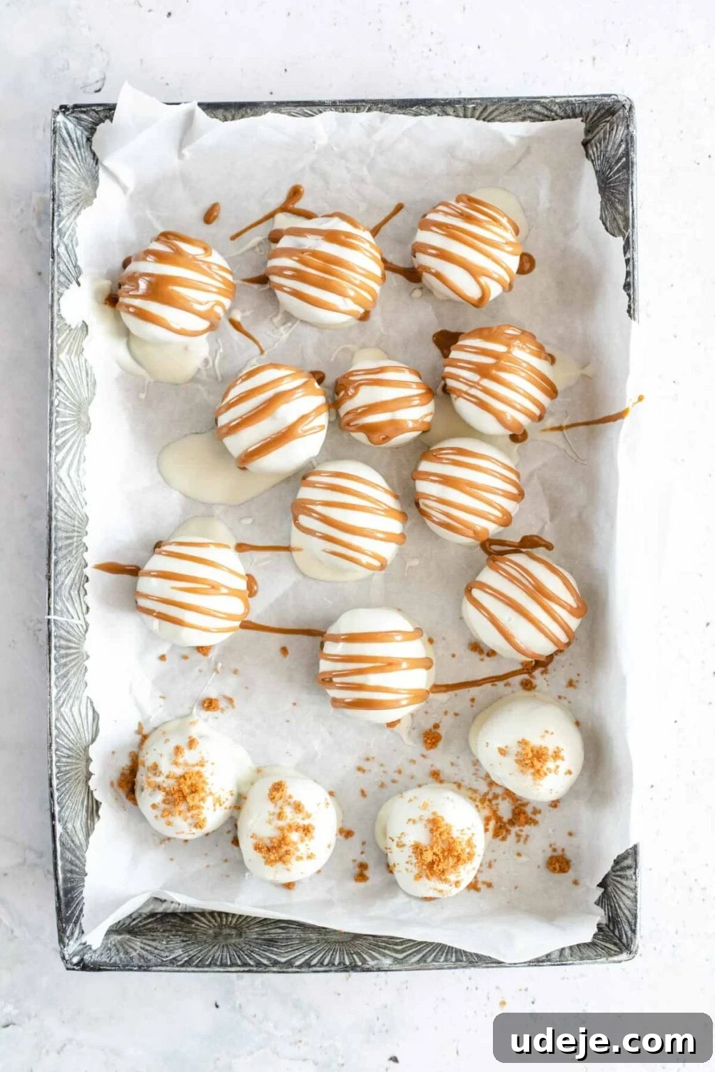 4-Ingredient No-Bake Biscoff Truffles 14 White chocolate covered Biscoff truffle balls, beautifully drizzled with melted Biscoff spread, arranged on parchment paper on a silver tray, set against a light marbled background.