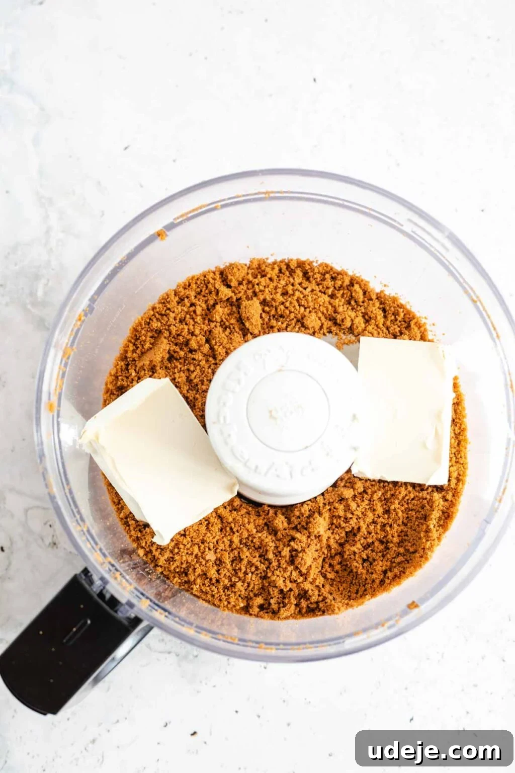 4-Ingredient No-Bake Biscoff Truffles 6 Biscoff cookie crumbs combined with softened cream cheese inside a food processor bowl, positioned on a light marbled background.