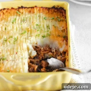A yellow baking dish filled with shepherd’s pie, reminiscent of a hearty potato moussaka, is topped with golden mashed potatoes and fresh herbs. A spoon rests in the dish, revealing the savory meat filling beneath.
