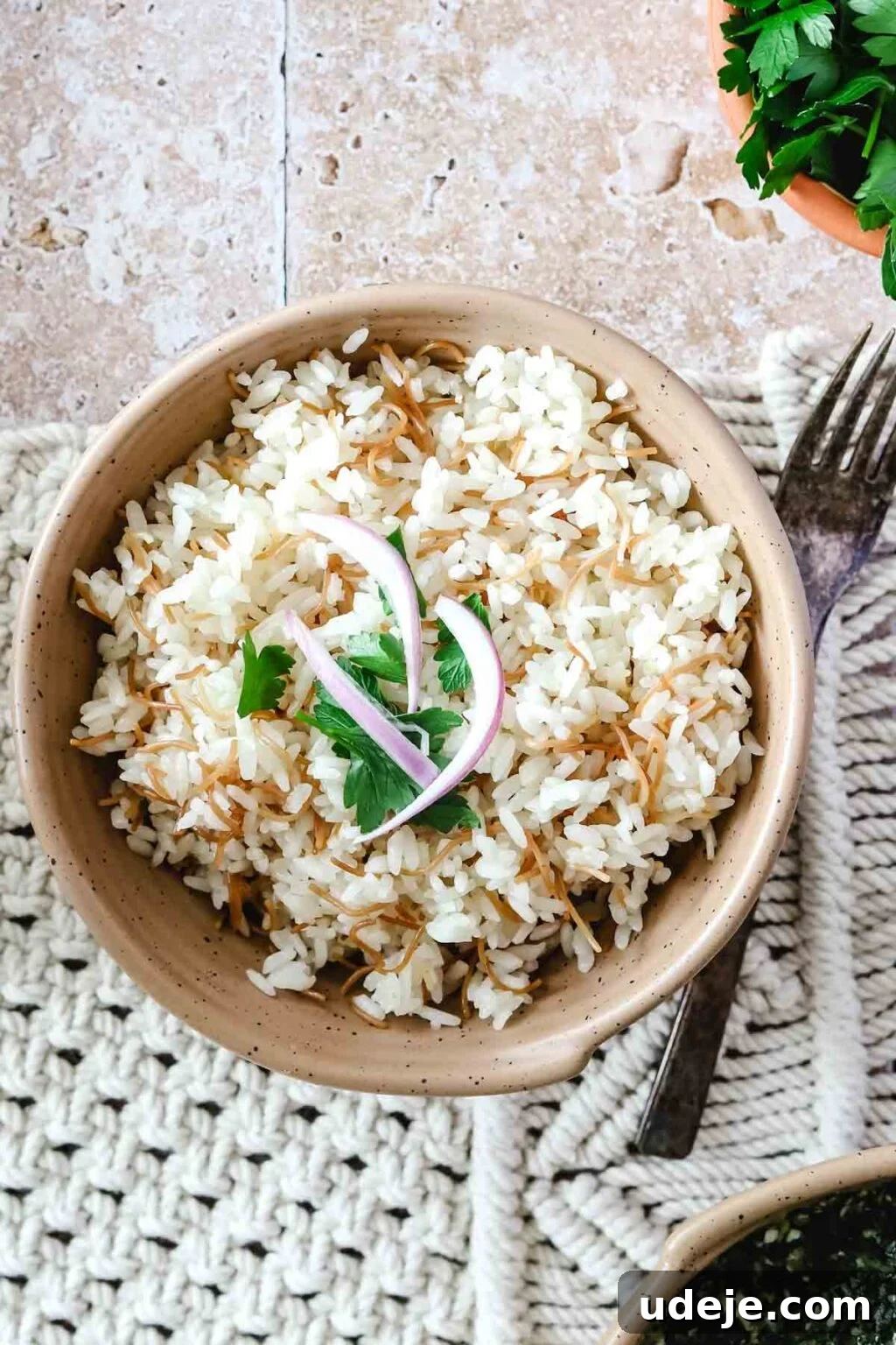 Egyptian Cloud Rice 2 Egyptian rice with vermicelli with sliced red onions and cilantro on top with a fork beside it on a woven background and a bowl of spinach and a bowl of cilantro on a pink tiled background