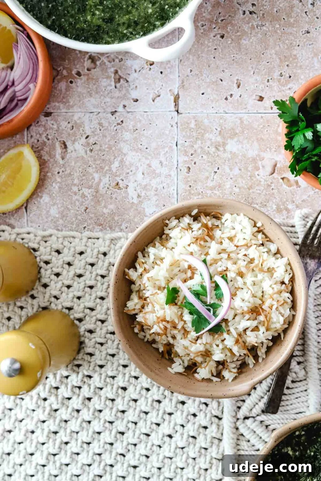 Egyptian Cloud Rice 10 Cooked Egyptian rice and vermicelli in a beige bowl with slices of red onion and cilantro on top on a woven background and a fork beside the bowl of rice, with salt and pepper shakers, wedge of lemon, bowl of onions and bowl of spinach and a bowl of cilantro on a pink tiled background