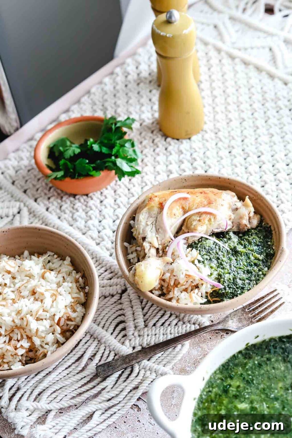 Molokhia: A Timeless Tradition 13 A beautifully plated beige bowl of Molokhia with rice, cooked chicken, and sliced red onions, a fork resting beside it on a woven mat. A separate white bowl of molokhia, rice in another beige bowl, and cilantro with salt and pepper shakers complete the inviting spread.