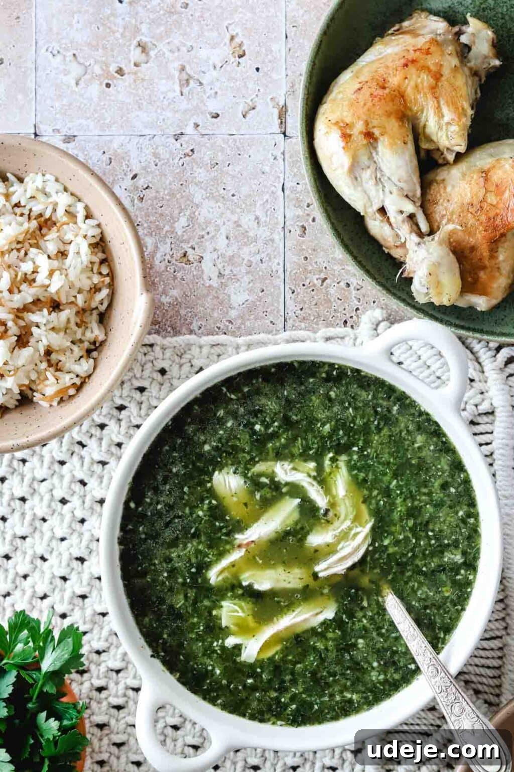 Molokhia: A Timeless Tradition 3 Molokhia in a white bowl with a metal spoon on a woven mat, accompanied by a bowl of rice in a beige bowl and cooked chicken on a green plate, all set against a pink tiled background.
