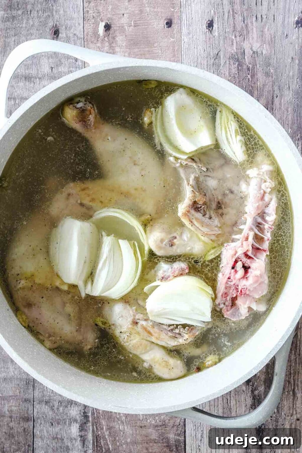 Molokhia: A Timeless Tradition 6 Large white pot filled with chicken pieces, simmering broth, and quartered onions on a wooden backdrop, illustrating the initial stage of broth preparation.