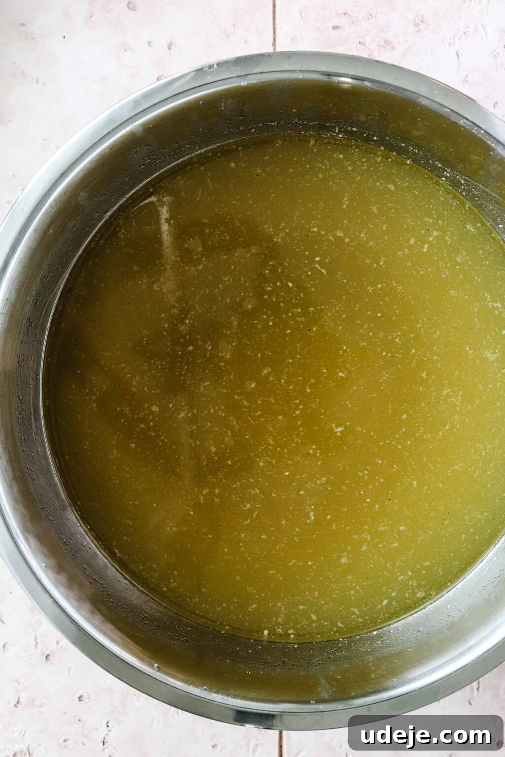 Molokhia: A Timeless Tradition 7 A glistening steel pot filled with crystal-clear, strained chicken broth, ready for the next step in preparing Molokhia, set on a charming pink tiled background.