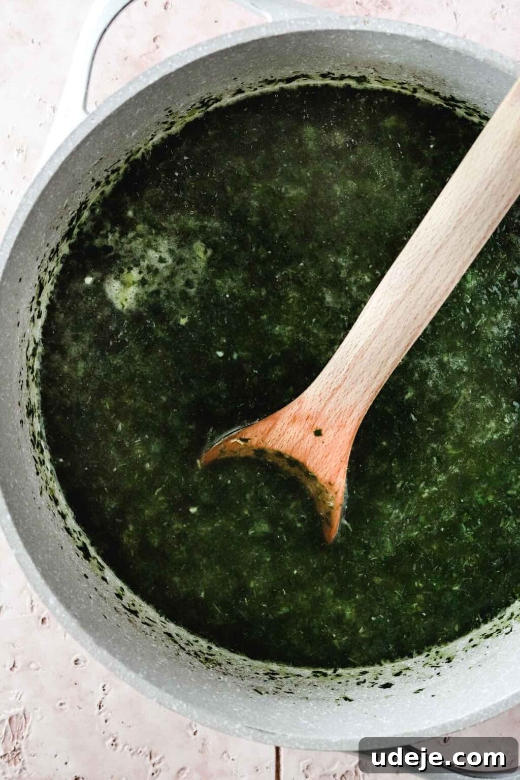 Molokhia: A Timeless Tradition 8 Molokhia leaves mixed with chicken broth, gently simmering in a large white pot with a wooden spoon, resting on a light pink tiled background.
