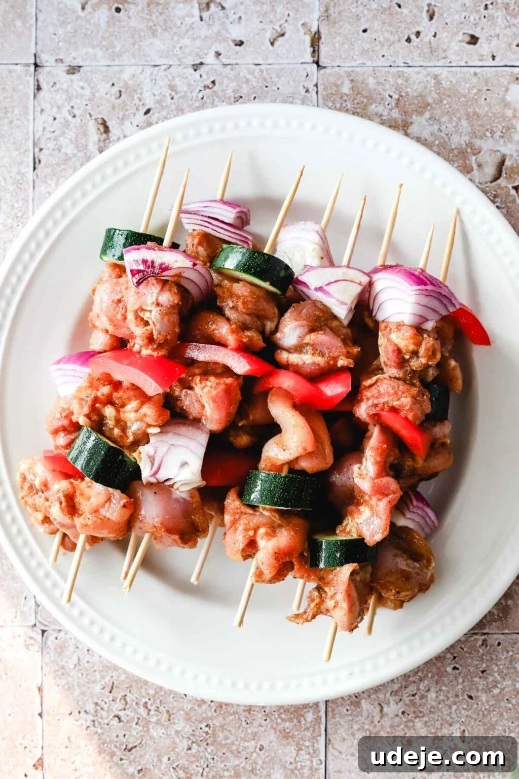 Effortless 15-Minute Air Fryer Chicken Skewers 11 Beautifully prepared raw chicken kabobs, artfully skewered with alternating pieces of zucchini, red onion, and red peppers on bamboo skewers, arranged on a crisp white plate on a stone tiled background, ready for air frying.