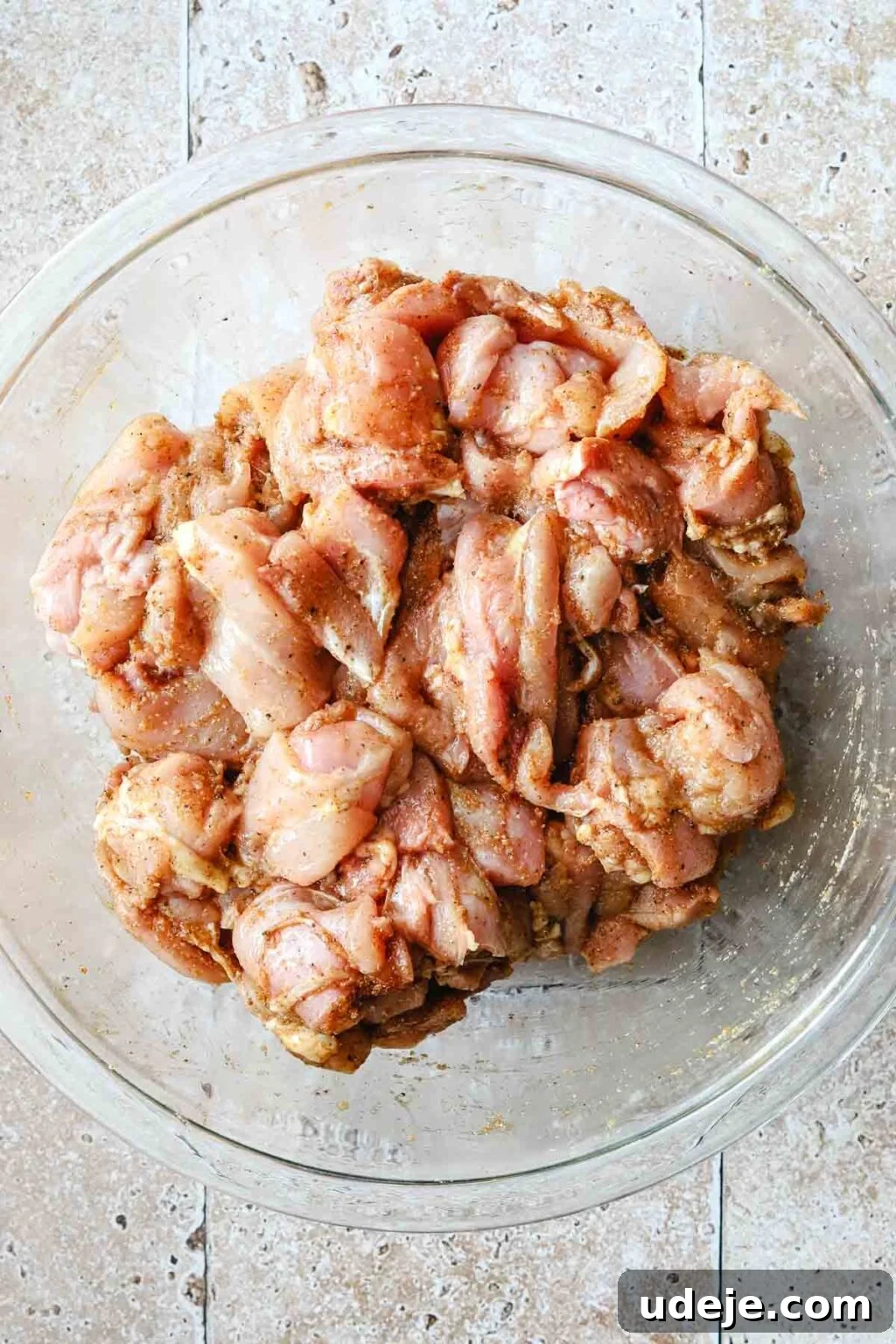 Effortless 15-Minute Air Fryer Chicken Skewers 12 Perfectly marinated boneless, skinless chicken thighs, glistening with spices and ready to be skewered, displayed in a glass bowl on a textured stone tiled background.