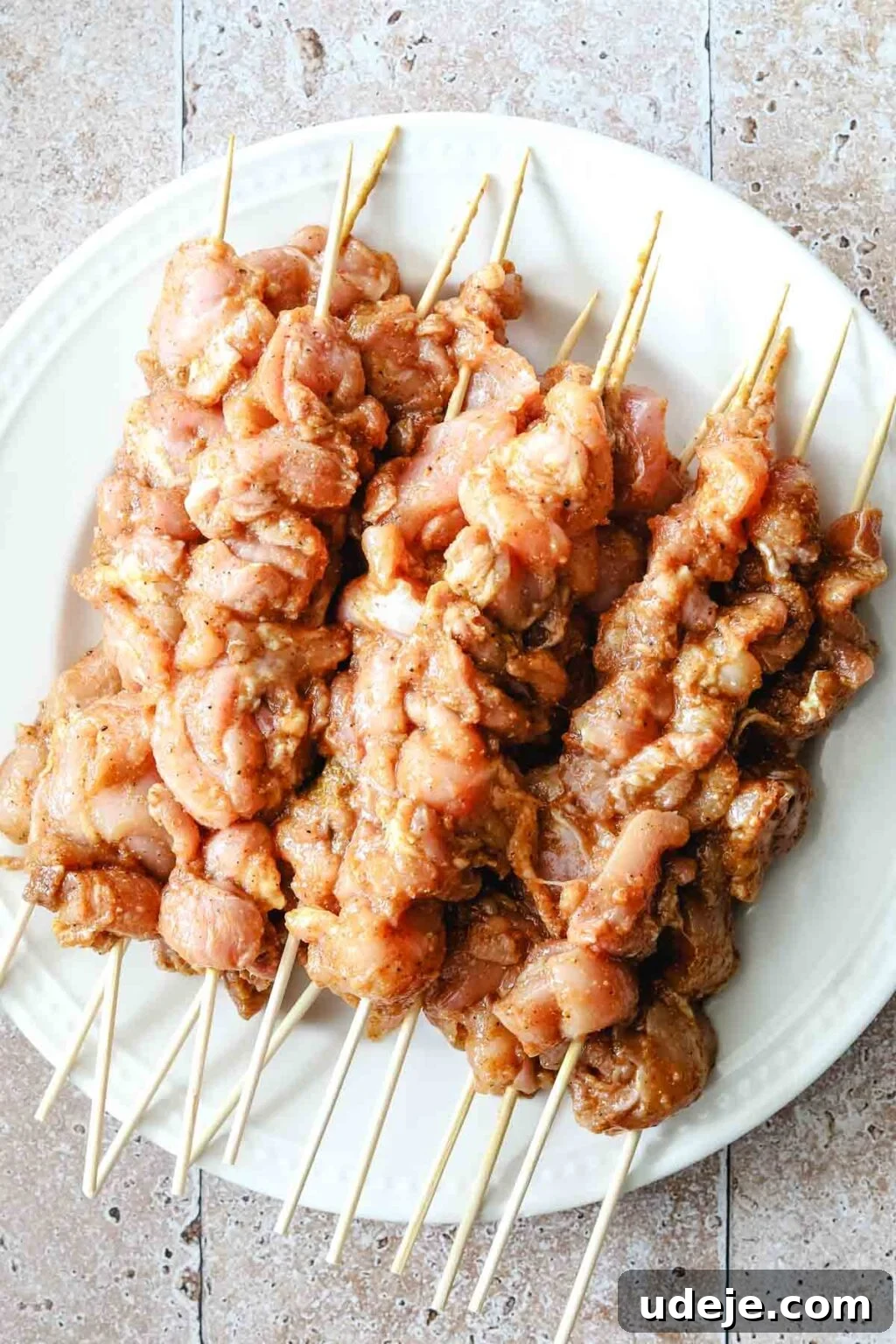Effortless 15-Minute Air Fryer Chicken Skewers 13 Raw, marinated chicken pieces neatly skewered on bamboo sticks, arranged on a pristine white plate atop a stone tiled background, awaiting their turn in the air fryer.