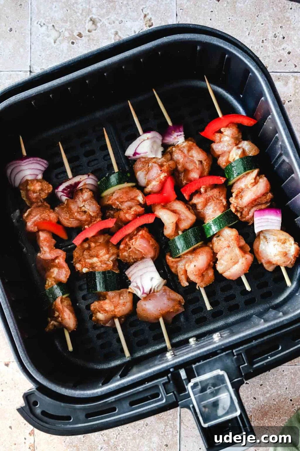 Effortless 15-Minute Air Fryer Chicken Skewers 14 Raw chicken kabobs, featuring zucchini, red onion, and red peppers on bamboo skewers, neatly arranged in a black air fryer basket on a stone tiled background, ready for cooking.