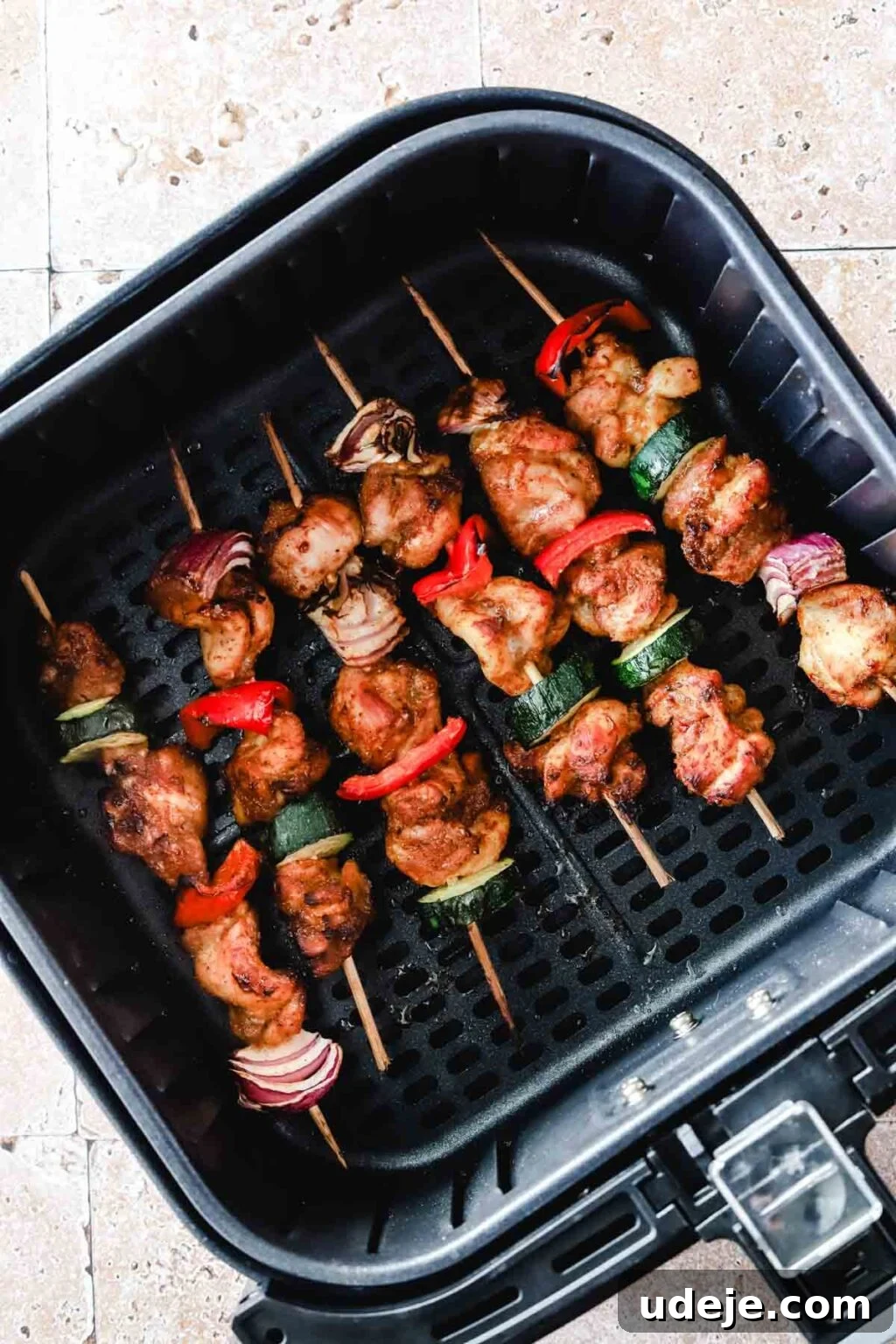 Effortless 15-Minute Air Fryer Chicken Skewers 15 Beautifully cooked chicken kabobs with tender zucchini, red onion, and red peppers on bamboo skewers, perfectly browned and resting in a black air fryer basket on a stone tiled background, ready to be served.