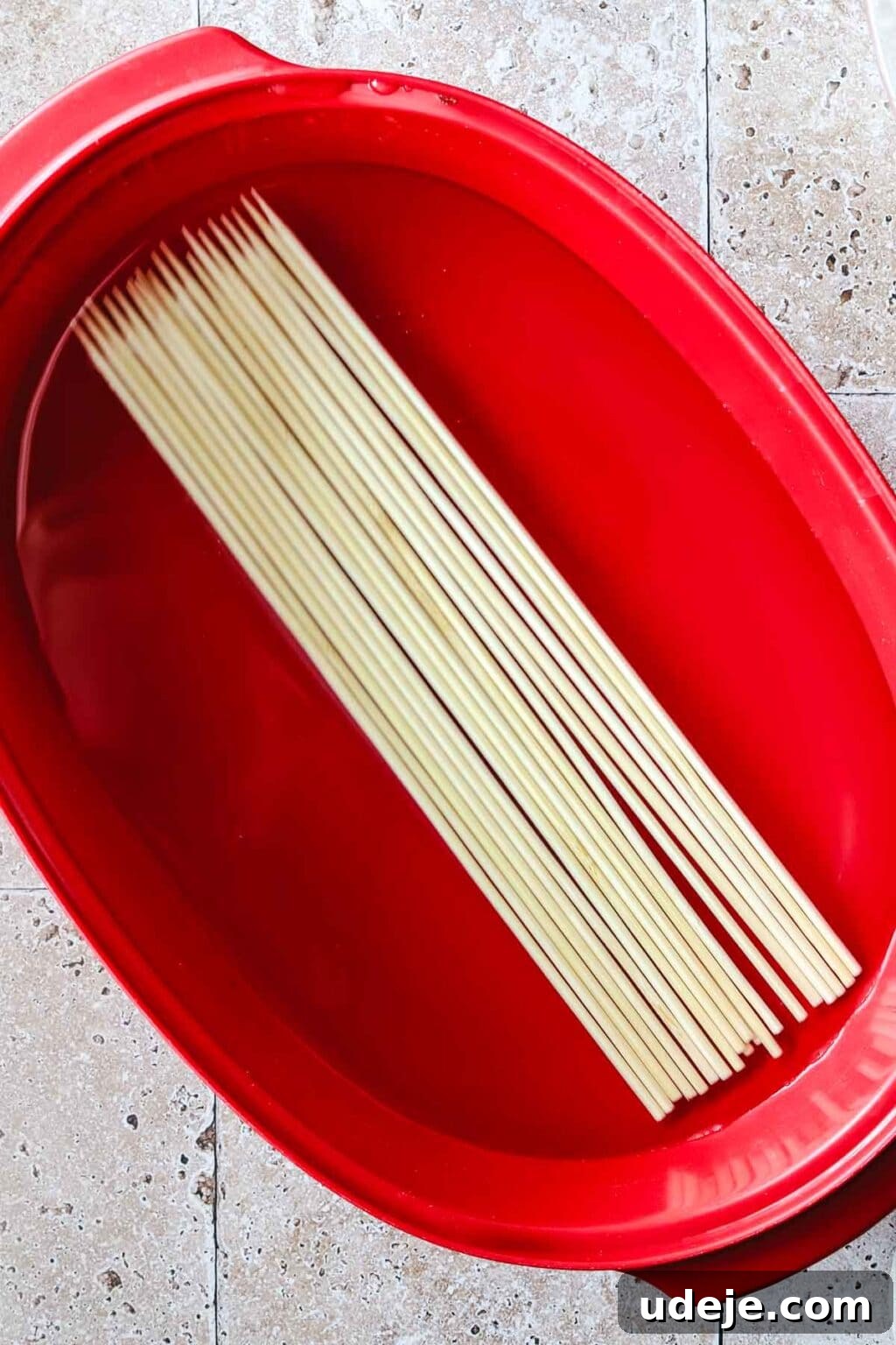 Effortless 15-Minute Air Fryer Chicken Skewers 8 Bamboo skewers gently soaking in water within a red oval dish, resting on a textured stone tiled background, preparing them for even cooking in the air fryer.