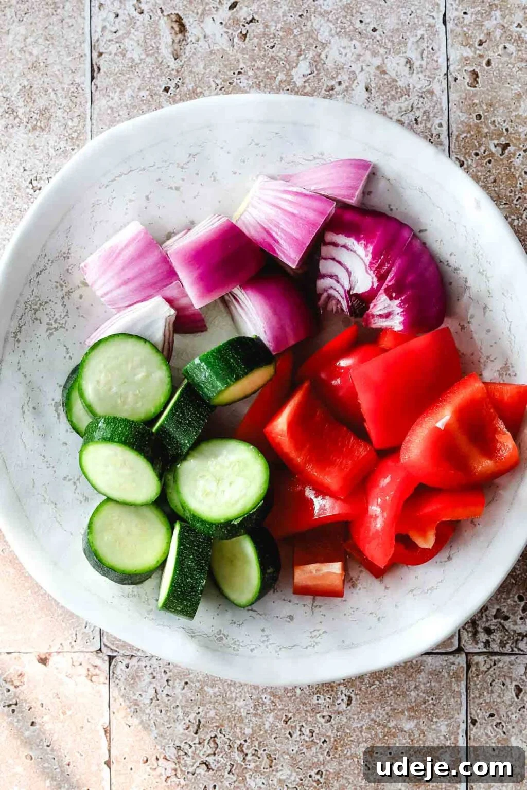 Effortless 15-Minute Air Fryer Chicken Skewers 10 A pristine white plate holds carefully cut pieces of red onion, zucchini, and red pepper, ready to be skewered alongside the chicken, all resting on a elegant stone tiled background.