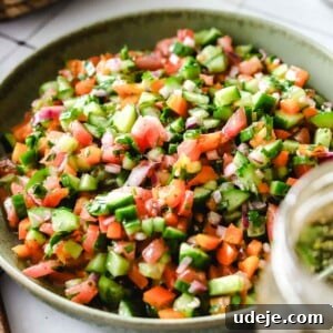 A finely chopped Arabic salad made up of cucumber, tomato, red pepper, red onions and parsley served in a shallow green bowl. A mason jar is blurred out in the foreground, and a basket of cucumbers is in the background.