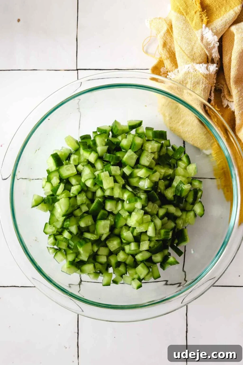 Effortless Arabic Salad 7 Finely chopped cucumber pieces are gathered in a medium glass bowl, set against a background with a yellow kitchen towel in the top right corner.