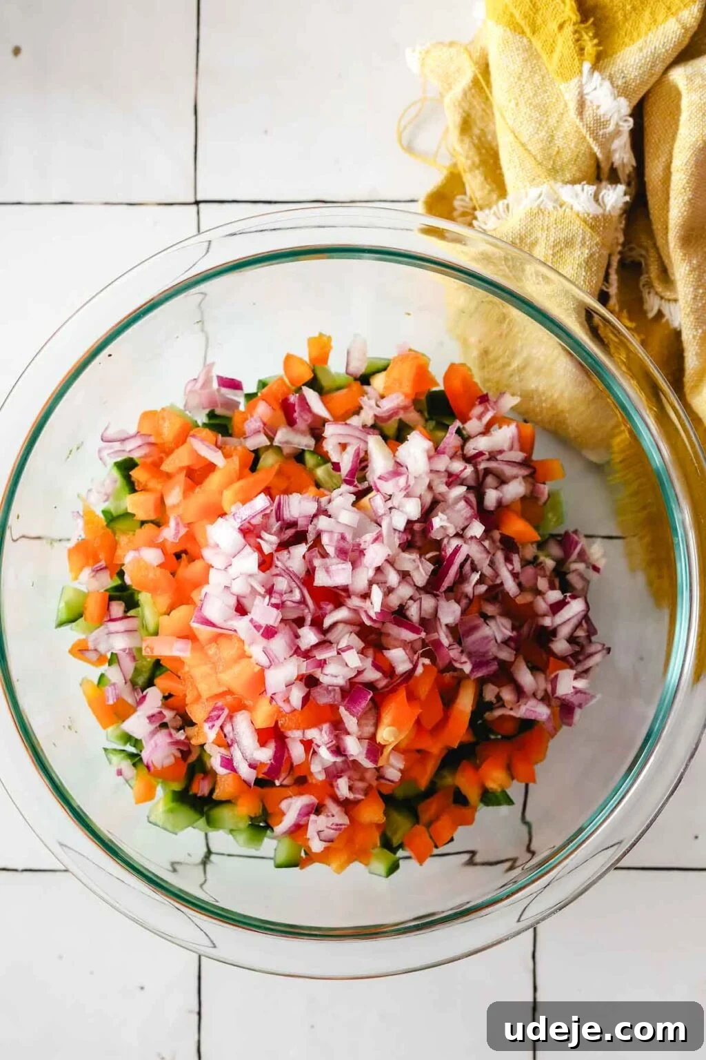 Effortless Arabic Salad 8 A mix of finely chopped cucumber, vibrant red bell pepper, and thinly sliced red onions fills a medium glass bowl, with a yellow kitchen towel adding a touch of color in the top right.