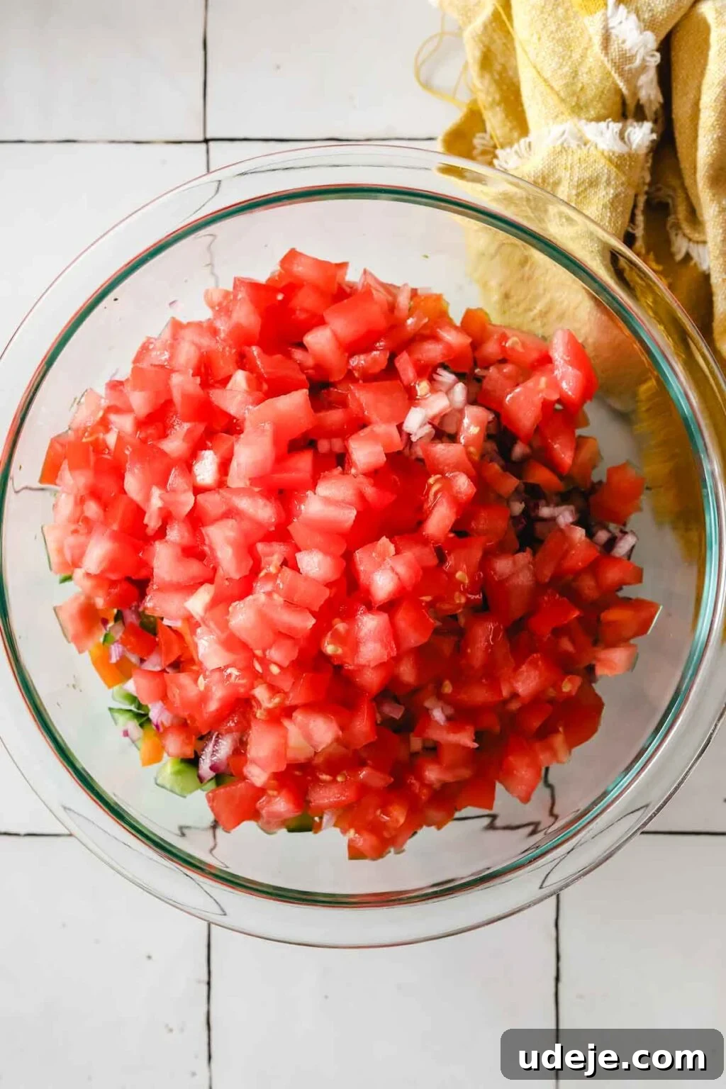 Effortless Arabic Salad 9 A colorful mixture of finely chopped cucumber, red bell pepper, ripe tomato, and red onions in a medium glass bowl, with a yellow kitchen towel corner visible.
