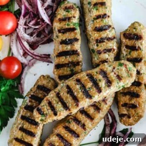 Grilled chicken kofta with grill marks on them, served on a white plate with sliced red onions, cherry tomatoes and parsley.