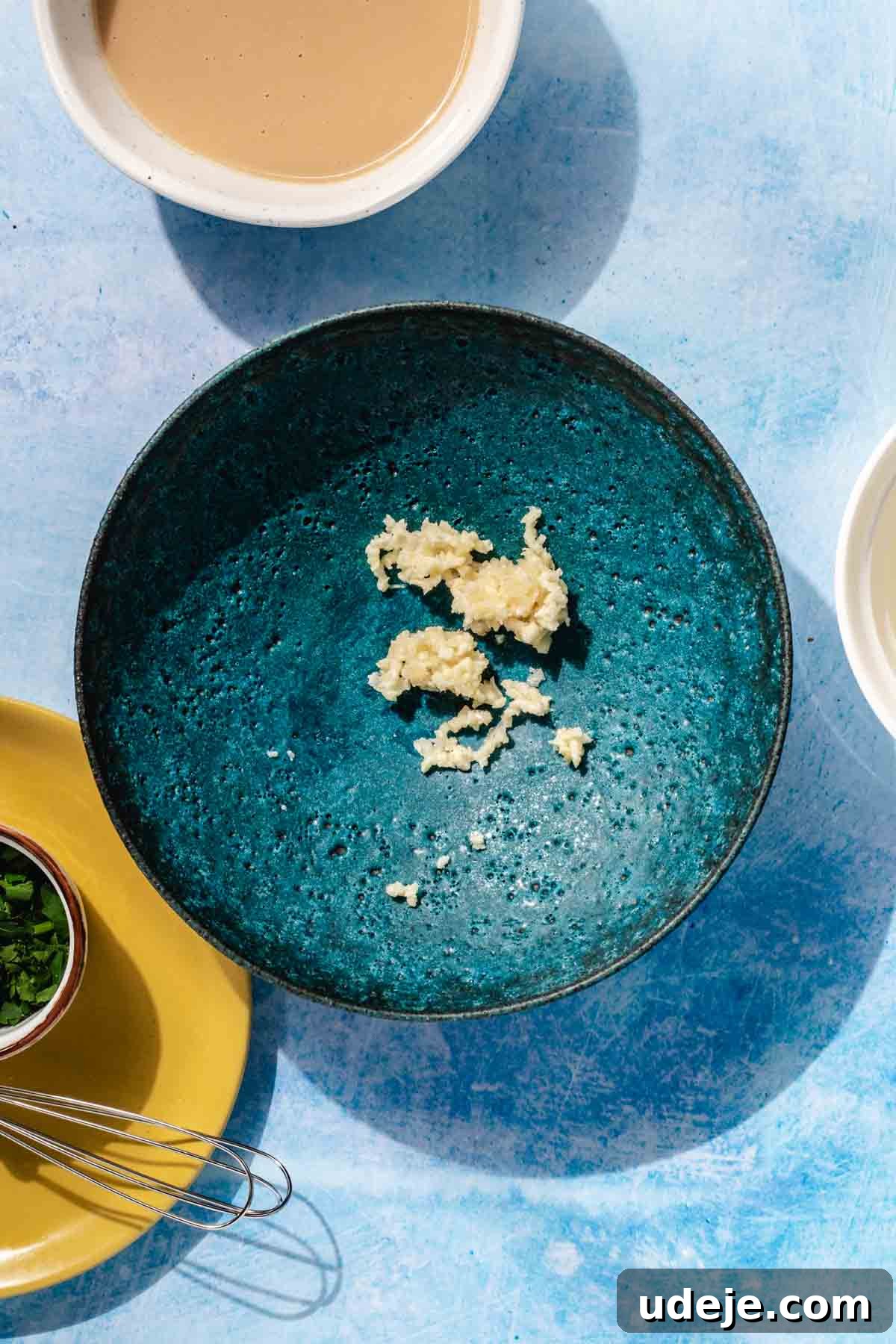 Simple & Creamy 3-Ingredient Tahini Sauce 4 Finely crushed garlic ready in a small blue stone bowl, with a larger bowl of tahini positioned to the side, along with fresh parsley and a small whisk, signaling the start of the sauce preparation.