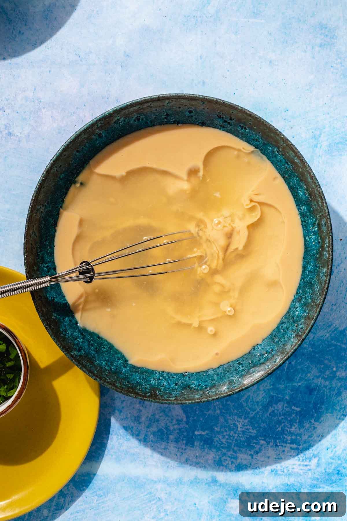 Simple & Creamy 3-Ingredient Tahini Sauce 6 A blue stone bowl containing crushed garlic, tahini, and water, with a small whisk submerged in the mixture, illustrating the initial stage of combining ingredients before whisking.