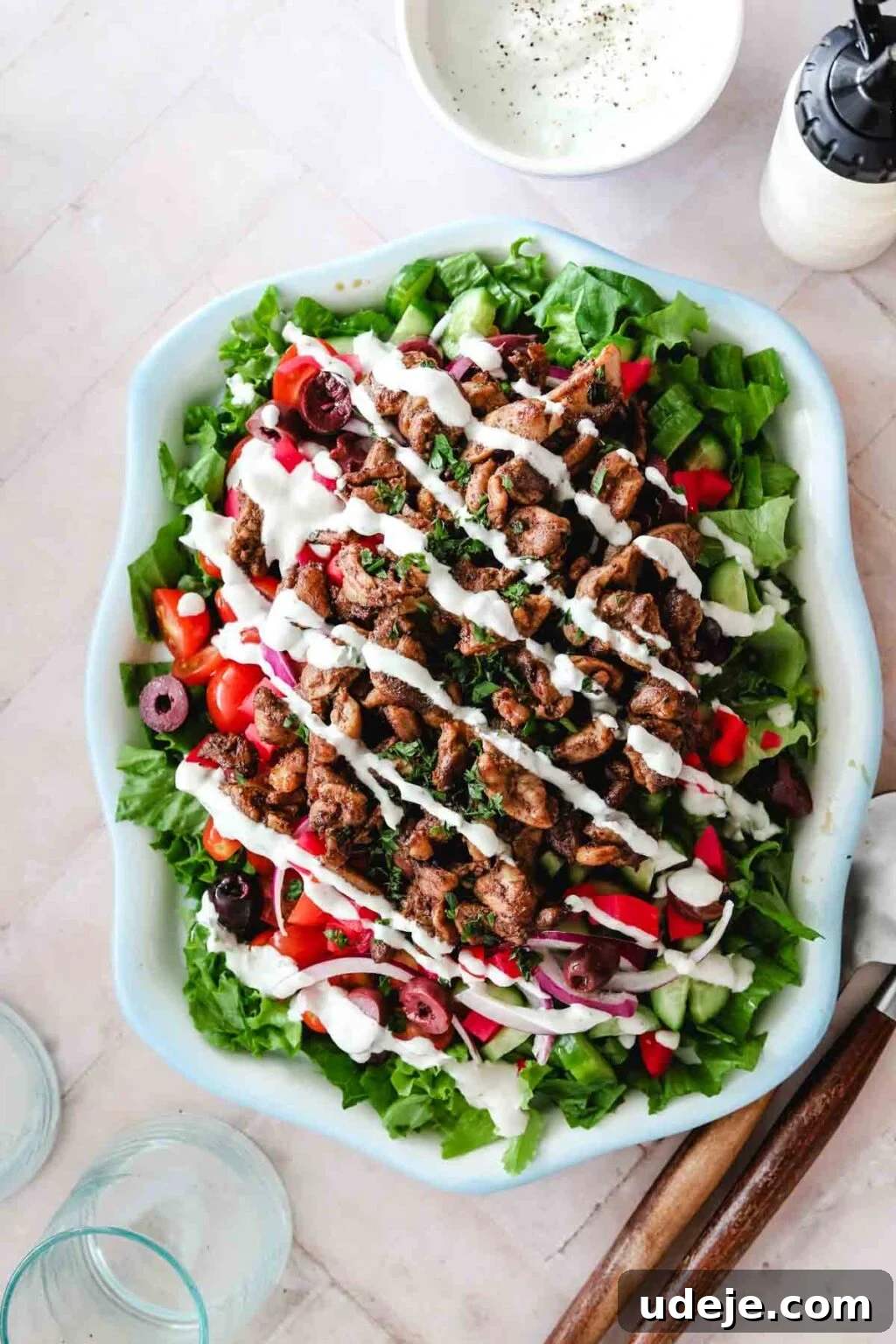 Creamy 5-Minute Shawarma Sauce 2 Shawarma salad in a light blue rimmed dish with a white bowl of shawarma sauce next to a squeeze bottle of shawarma sauce, with blue glasses in the bottom corner and wooden salad servers at the side on a light pink tiled background