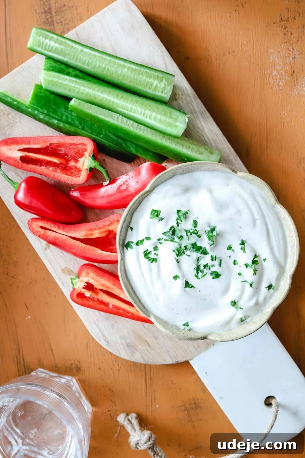 Creamy 5-Minute Shawarma Sauce 3 White bowl with shawarma sauce with herbs on it on a cutting board with cucumber sticks and mini red peppers, on an orange background a glass of water at the side