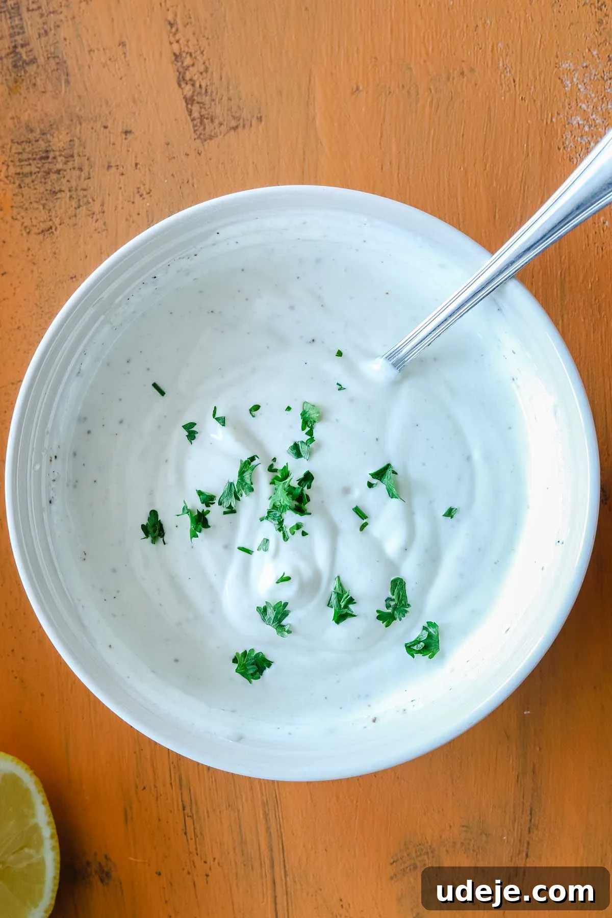 Creamy 5-Minute Shawarma Sauce 6 white bowl with shawarma sauce and herbs on top and a spoon in it on an orange background with a half lemon at the side