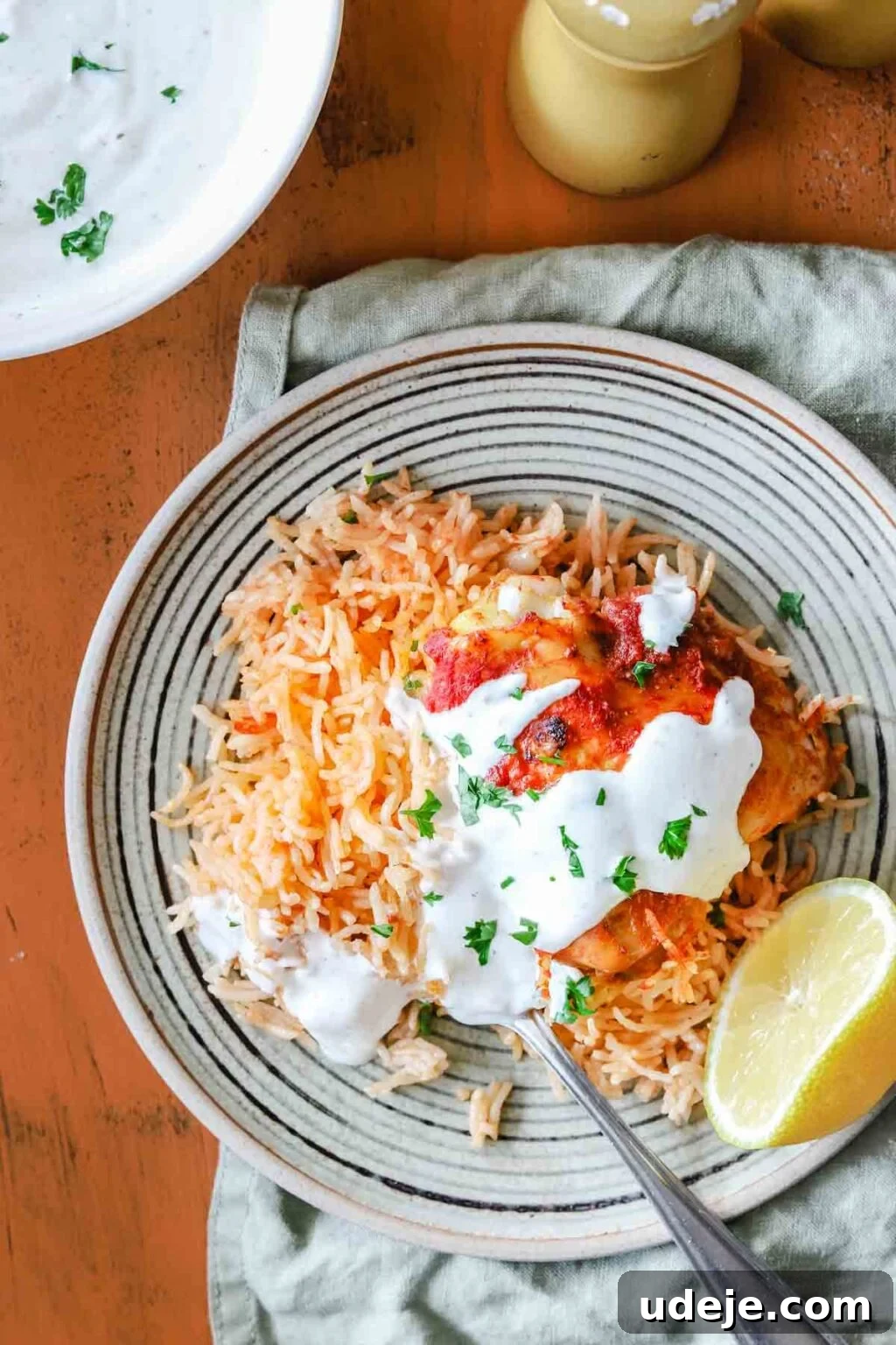 Creamy 5-Minute Shawarma Sauce 7 Plate of reddish rice on a green striped plate with shawarma sauce on it with a spoon in it and a half a lemon at the side on a green tea towel with a white bowl of shawarma sauce, pepper mill on an orange background