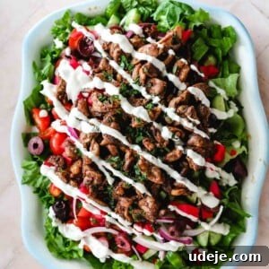 Lettuce, chopped cucumbers, sliced red onions, chopped pickled turnips, sliced black olives and halved cherry tomatoes, topped with chicken shawarma pieces and drizzled with shawarma sauce in a blue rimmed dish on a light pink tiled background