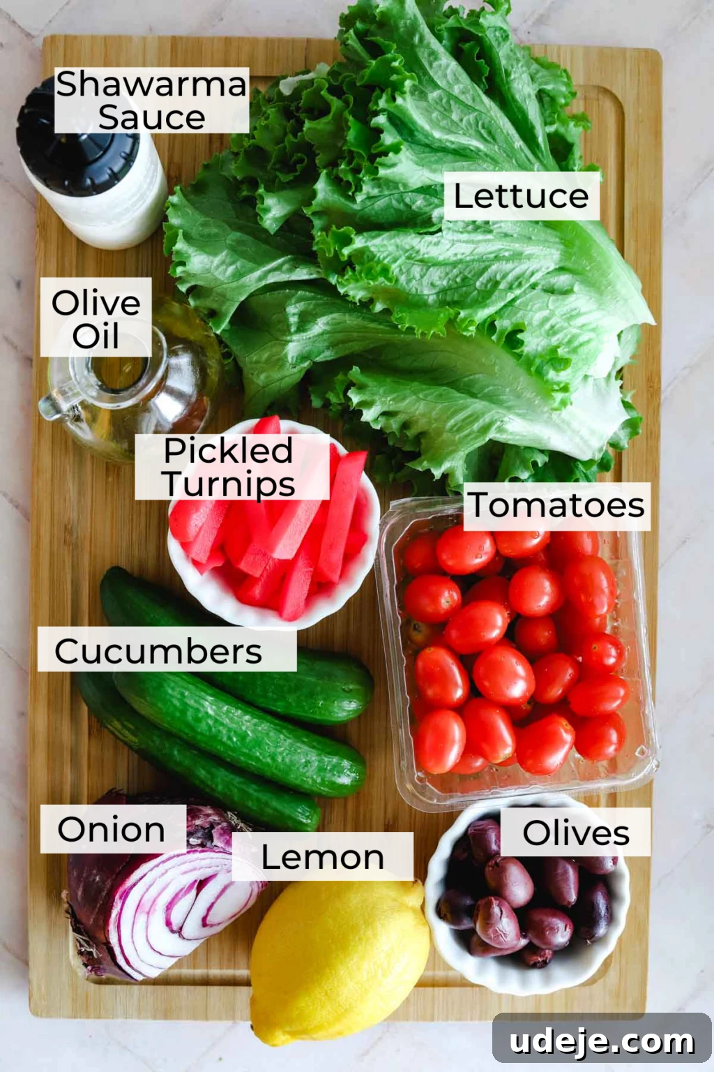 A beautiful array of fresh ingredients for a Shawarma Salad on a wooden cutting board, including romaine lettuce leaves, a bottle of olive oil, a bowl of pink pickled turnips, vibrant cherry tomatoes, crisp baby cucumbers, a red onion, a lemon, and black olives in a small white bowl. All essential for creating a healthy Middle Eastern salad.