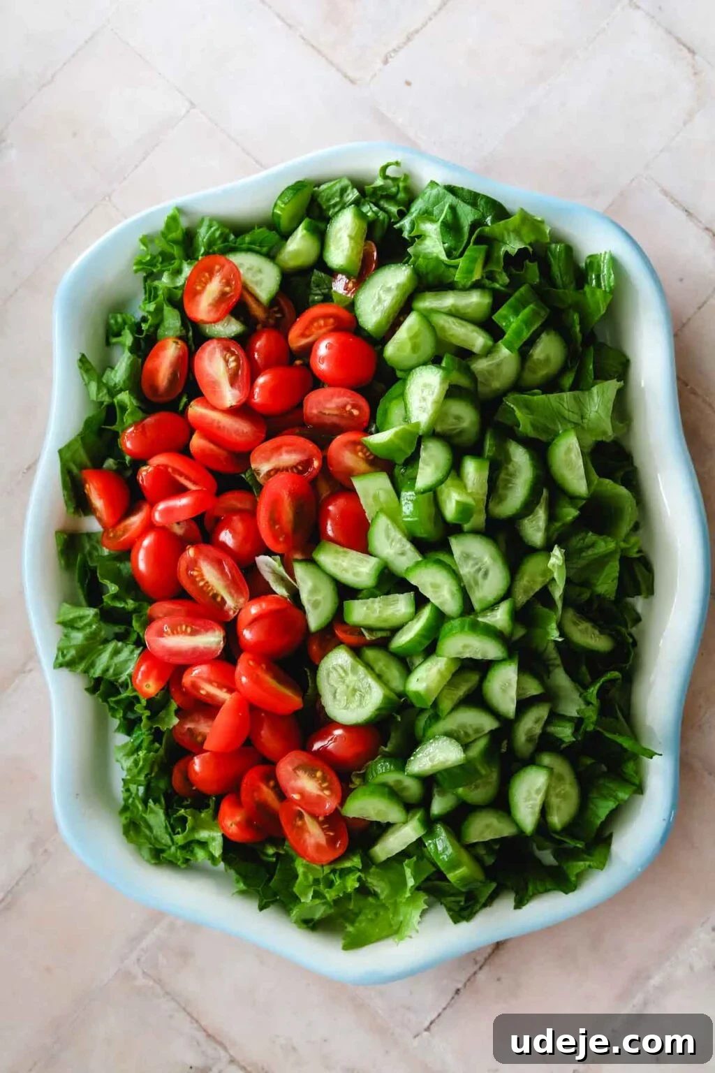 A partially assembled salad in a blue-rimmed dish, featuring a base of crisp lettuce, chopped cucumbers, and halved cherry tomatoes on a light pink tiled background, ready for more delicious additions.