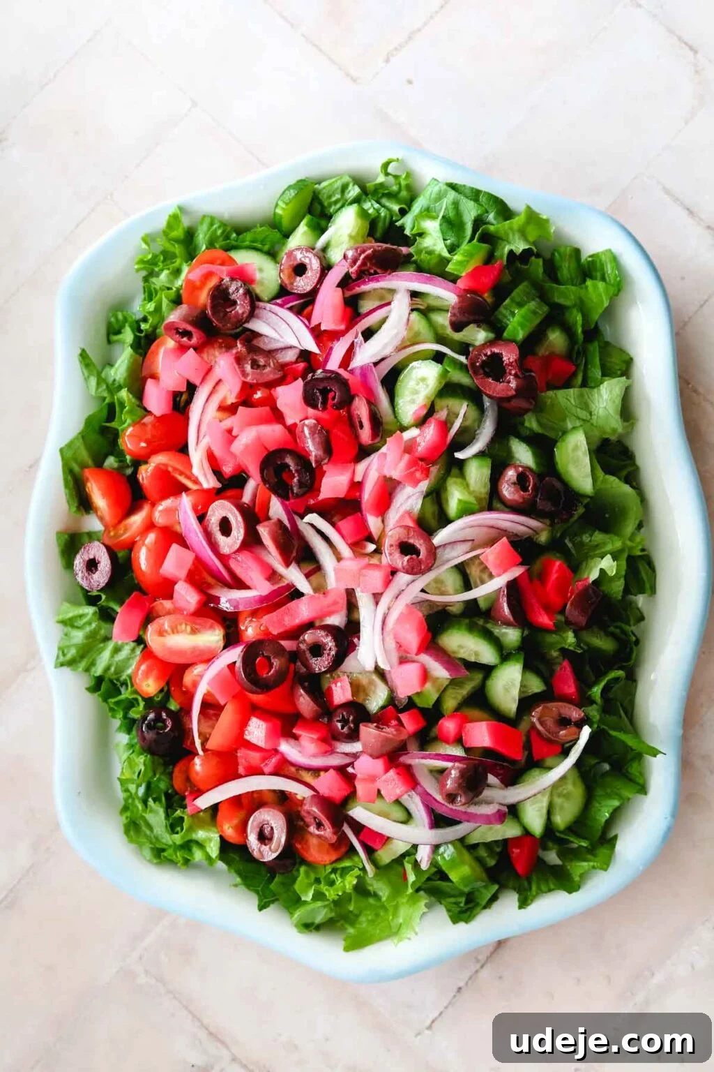 The salad continues to build in a blue-rimmed dish with added thinly sliced red onions, finely chopped pickled turnips, and sliced black olives, complementing the lettuce, cucumbers, and cherry tomatoes on a light pink tiled background.