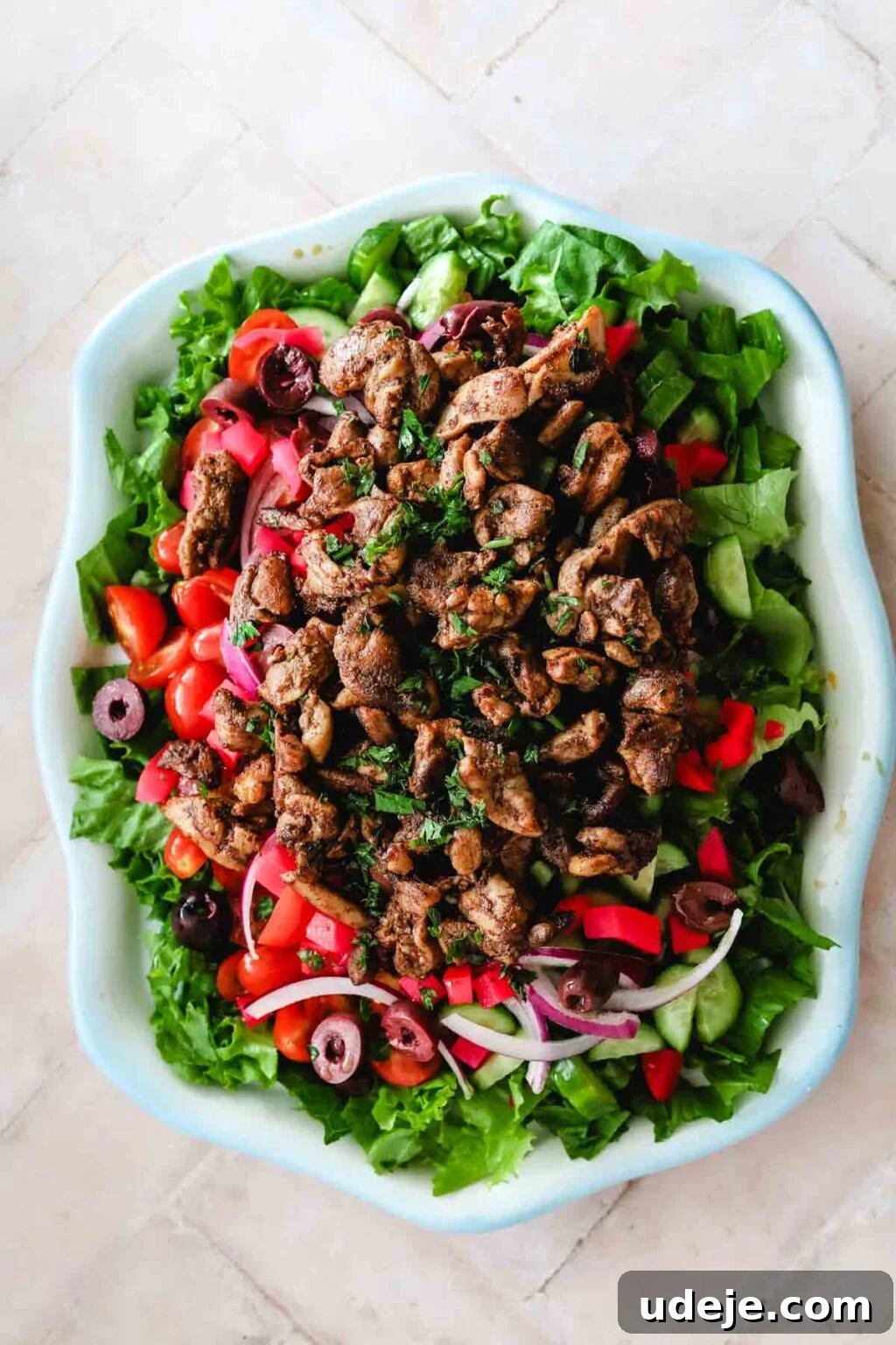 A fully assembled Chicken Shawarma Salad in a blue-rimmed dish, with a base of lettuce, cucumbers, tomatoes, red onions, pickled turnips, and black olives, magnificently topped with warm chicken shawarma pieces on a light pink tiled background.