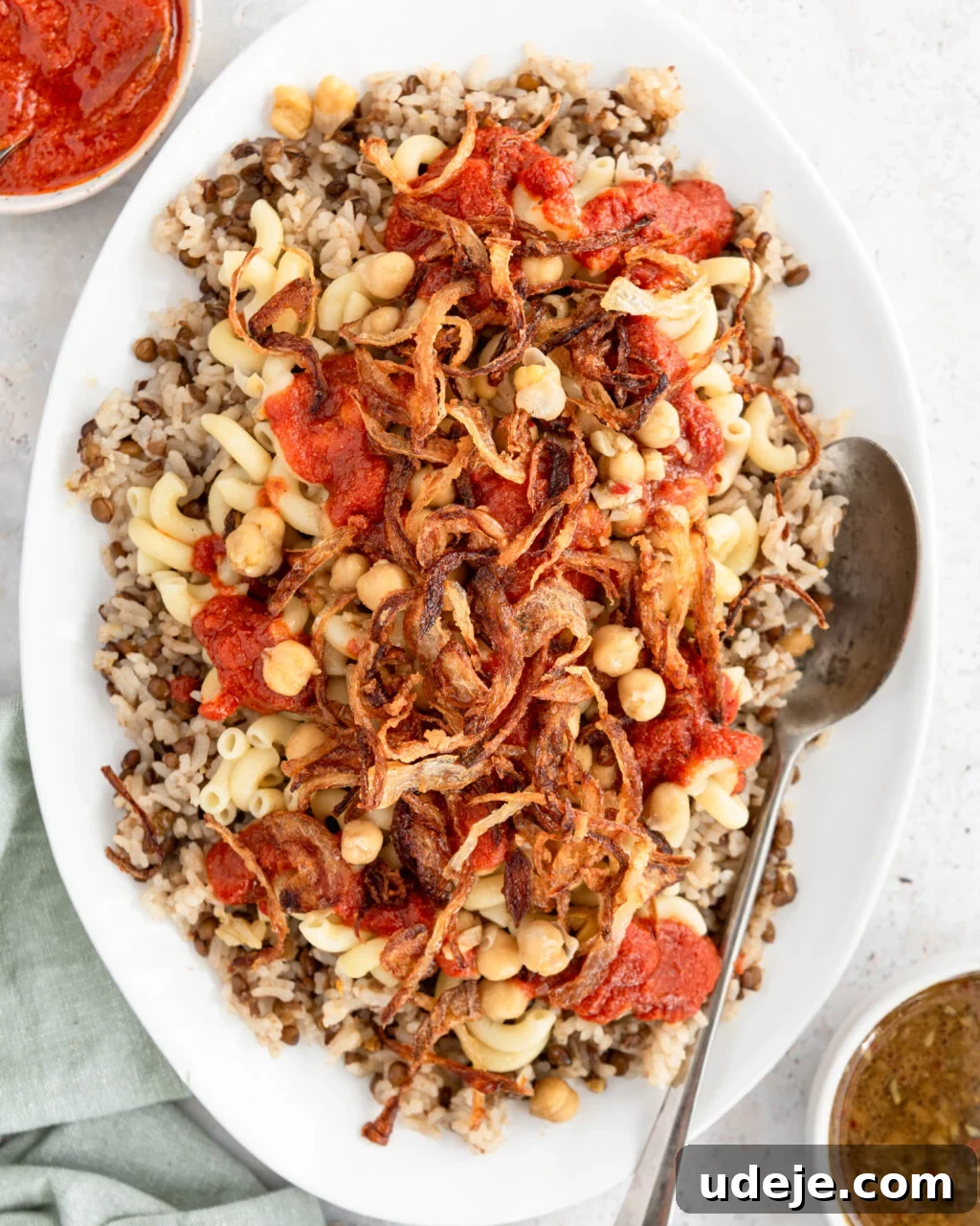 Egypt's Koshari: A Hearty Blend of Rice, Lentils and Pasta 2 Cooked rice and lentils with macaroni, spiced chickpeas, fried onions and tomato sauce on top on a white platter with a green towel at the side and a serving spoon, a bowl of tomato sauce and a bowl of dukkah on a light coloured background