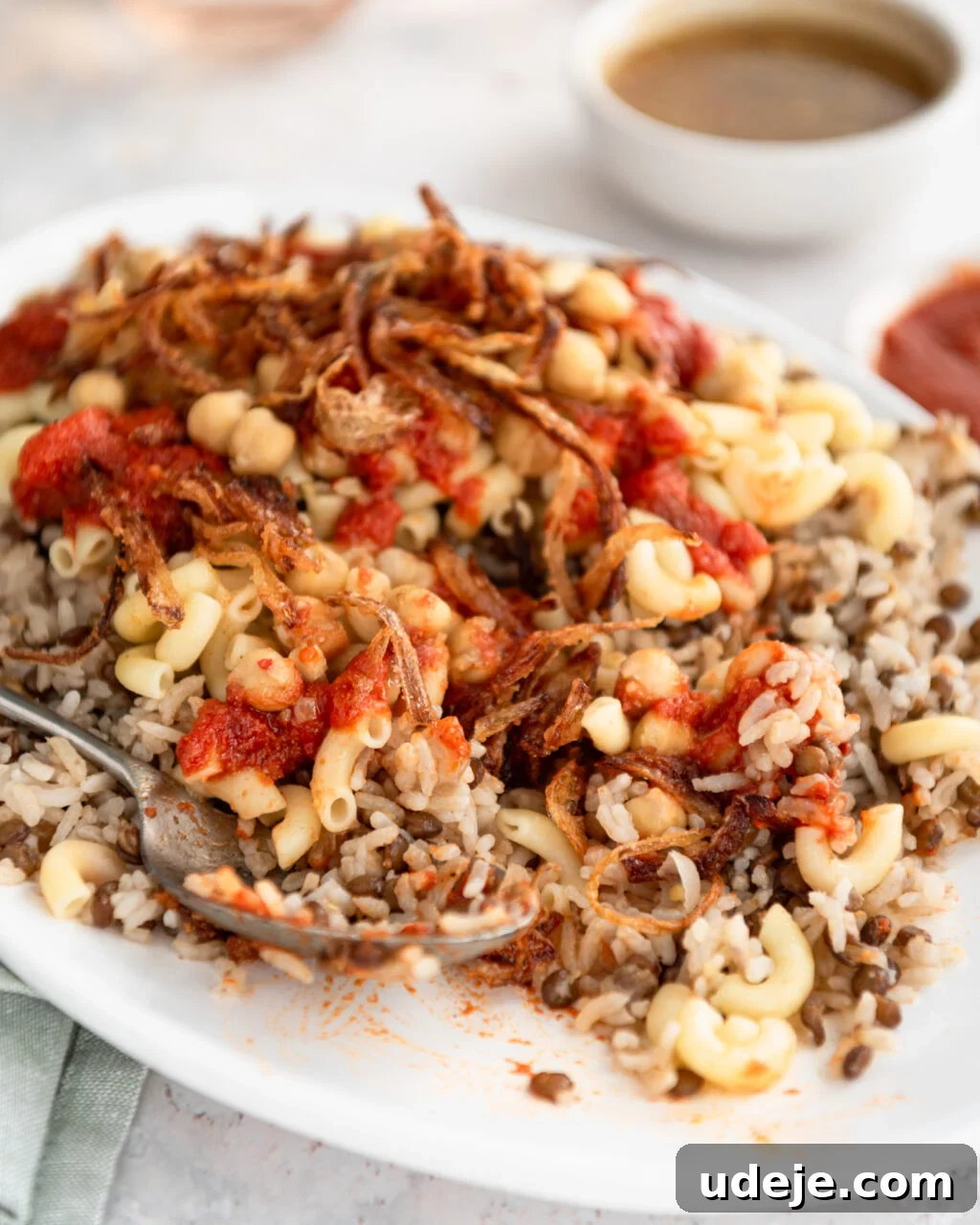 Egypt's Koshari: A Hearty Blend of Rice, Lentils and Pasta 3 Cooked rice and lentils with macaroni, spiced chickpeas, fried onions and tomato sauce on top on a white platter with a green towel at the side and a serving spoon, a bowl of tomato sauce and a bowl of dukkah and glasses of water at the back on a light coloured background