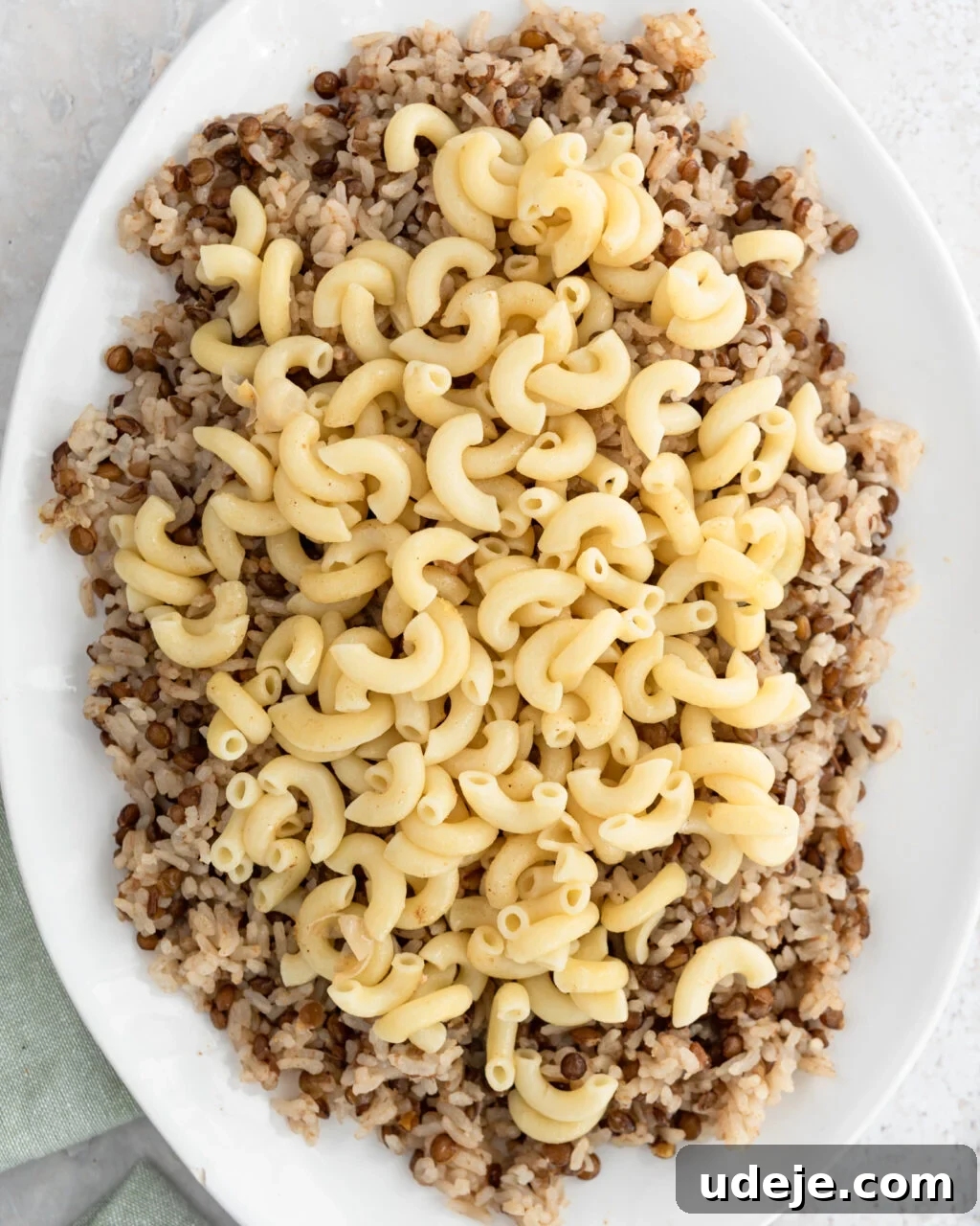 Egypt's Koshari: A Hearty Blend of Rice, Lentils and Pasta 21 Cooked rice and lentils with macaroni on top on a white platter with a green towel at the side on a light coloured background