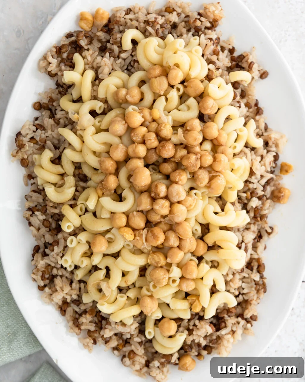 Egypt's Koshari: A Hearty Blend of Rice, Lentils and Pasta 22 Cooked rice and lentils with macaroni and spiced chickpeas on top on a white platter with a green towel at the side on a light coloured background