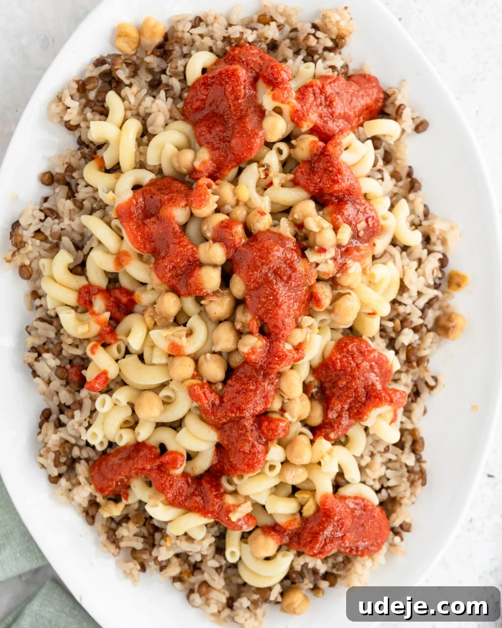 Egypt's Koshari: A Hearty Blend of Rice, Lentils and Pasta 23 Cooked rice and lentils with macaroni, spiced chickpeas and tomato sauce on top on a white platter with a green towel at the side on a light coloured background