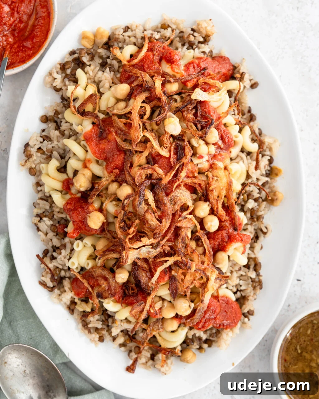 Egypt's Koshari: A Hearty Blend of Rice, Lentils and Pasta 24 Cooked rice and lentils with macaroni, spiced chickpeas, fried onions and tomato sauce on top on a white platter with a green towel at the side and a serving spoon, a bowl of tomato sauce and a bowl of dukkah on a light coloured background